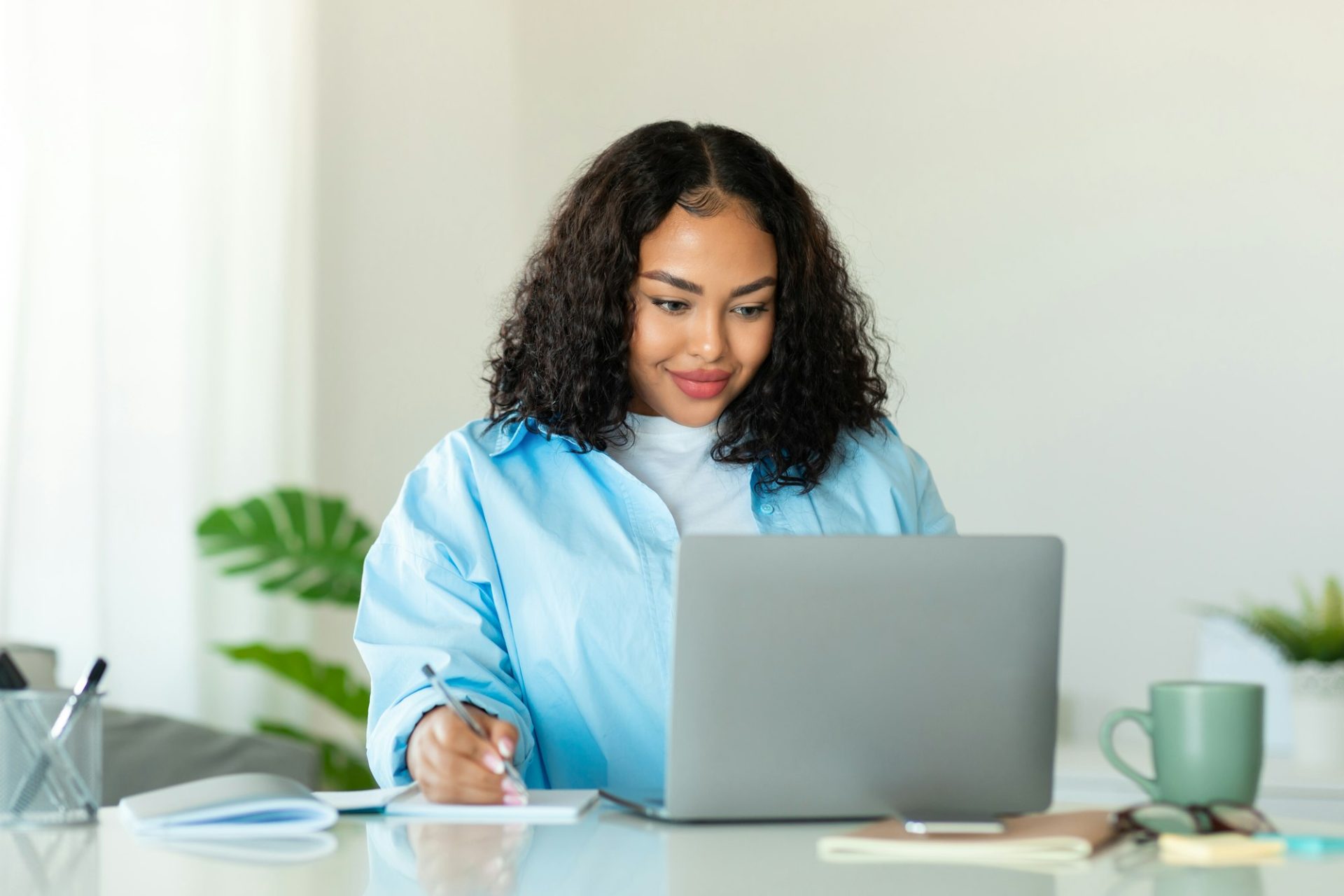 black-woman-sitting-at-desk-using-laptop-and-writing-in-notebook